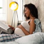 Pregnancy And Breast Implants - Pregnant woman resting on a bed while reading a book.