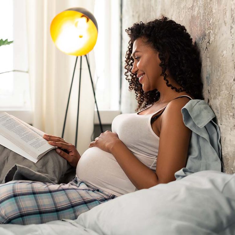Pregnancy And Breast Implants - Pregnant woman resting on a bed while reading a book.